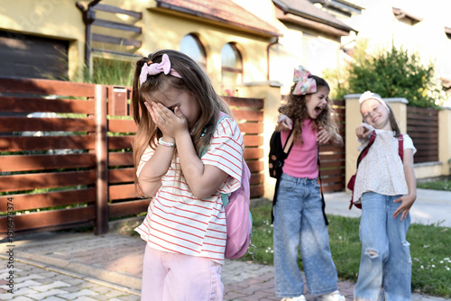 Two primary school girls laughing and pointing fingers at a sad classmate covering her face outdoors; concept of school bullying and peer pressure