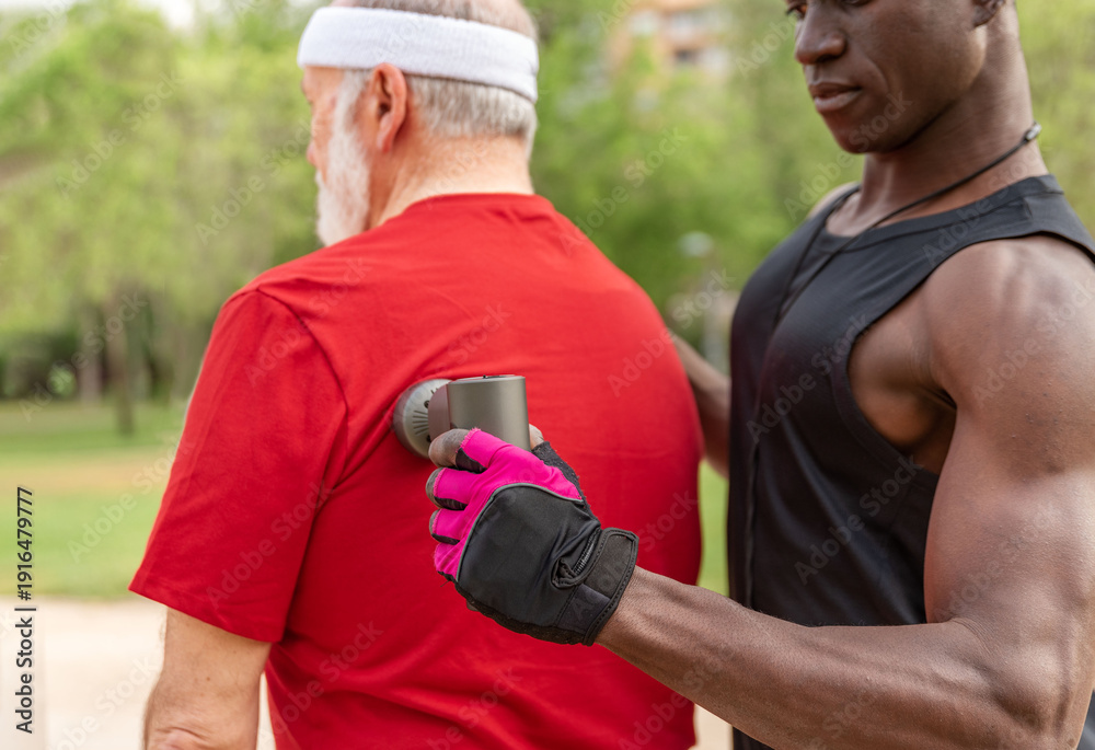 Naklejka premium Close up of Strong personal trainer giving massage therapy with gun to athletic elderly man outdoors