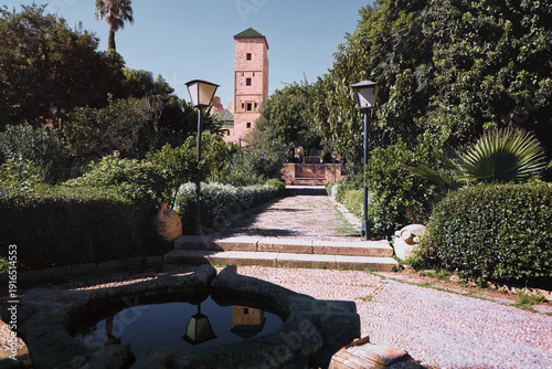 A scenic pathway bordered by greenery leads to a historic stone tower under a clear sky, evoking a serene and tranquil atmosphere.