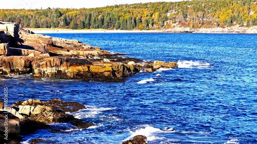 Scenic view of the Atlantic ocean crashing on the rocky coast of Maine. Beautiful autumn foliage covering the landscape on a sunny day with Sandy Beach in distance.