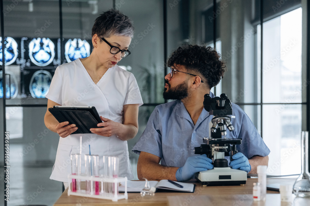 Fototapeta premium Female healthcare professional in white uniform discusses findings with male colleague in blue scrubs at laboratory table with microscope and test tubes in modern medical facility