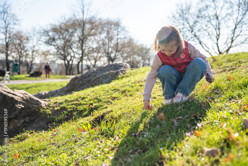 Little girl exploring nature in city park during spring walk. Curious child interacting with soil, leaves and landscape in outdoor environment.