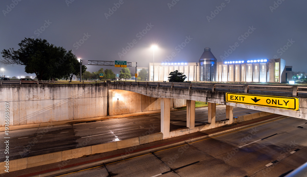 Obraz premium Oklahoma History Center in Oklahoma City. Modern museum building features blue illuminated lettering and a glass atrium behind a highway overpass with an exit sign on a foggy night