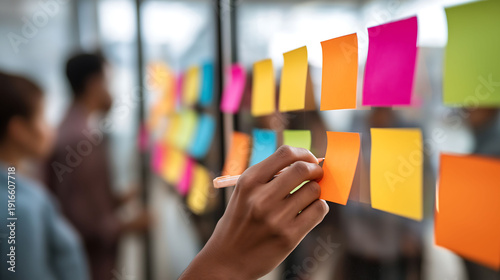 A hand places a colorful sticky note on a glass board during a team meeting, representing brainstorming, collaboration, and creative project planning in a modern office