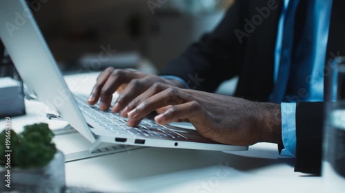 Close up male hands typing on laptop keyboard while working late at office desk with business documents nearby. Focused professional finishing tasks on computer in modern workspace during evening.