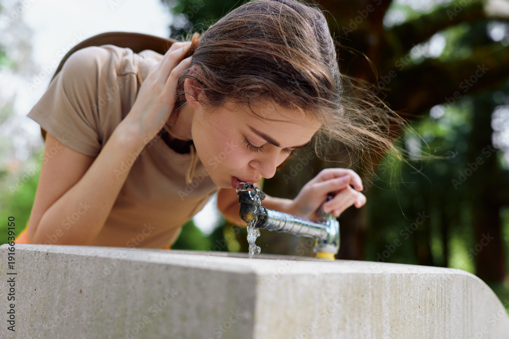 Fototapeta premium Woman drinking from a public fountain in a park, young female leaning over a stone basin to sip water, candid portrait of a refreshing hydration moment with green trees and sunlight