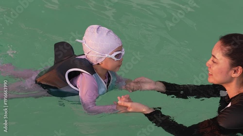 slow motion of preschool child girl in life jacket learning to swim with her mother in swimming pool