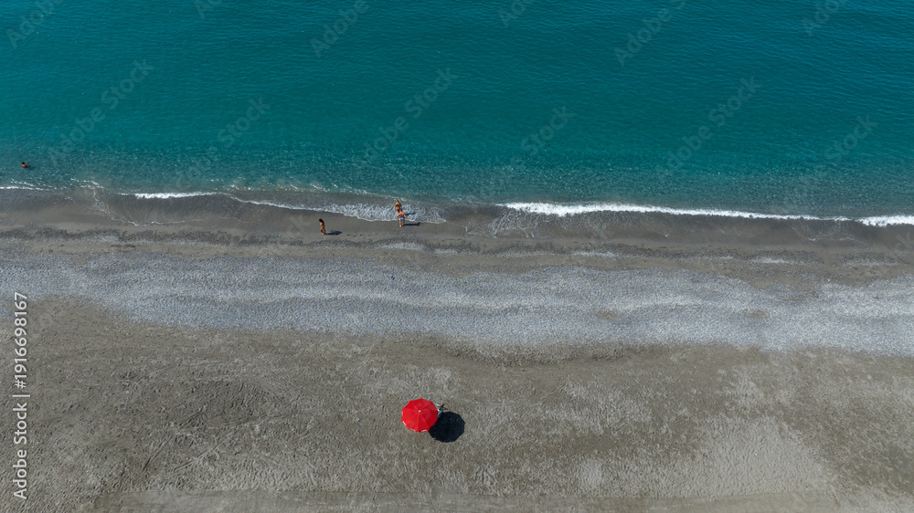 Obraz premium Minimalist aerial view of a wide pebble beach where a single red umbrella stands as a vibrant focal point. In the distance, near the shoreline, two small figures walk by the turquoise waves of the sea