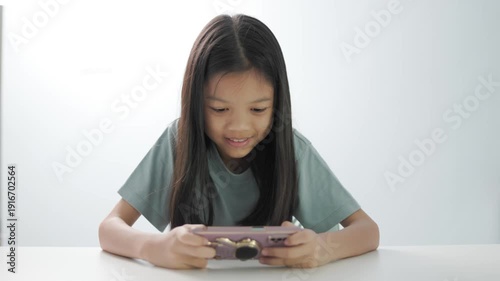 Young Asian girl using smartphone on white table. Concept of online education, digital literacy for kids, mobile gaming, and social media addiction. Isolated on bright background with copy space.