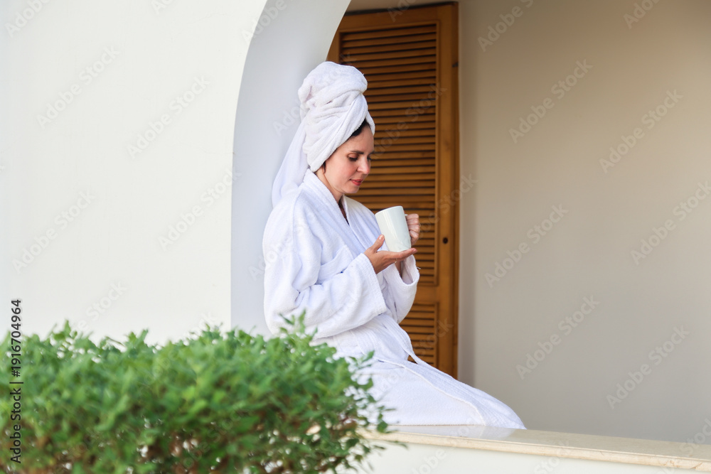 Fototapeta premium A woman in a robe and towel drinks a hot drink a terrace after a shower.