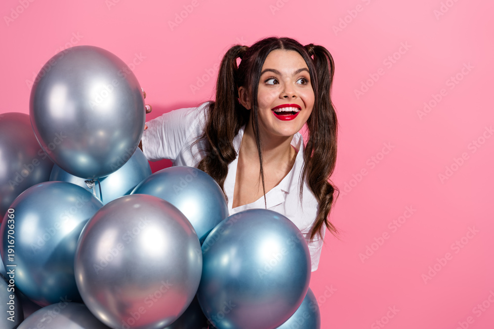 Fototapeta premium Joyful young woman with blue balloons poses against pink background smiling brightly in a casual summer fashion shoot