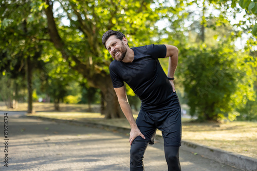 Fotografie A young male athlete stands in the park, bent over due to back pain, holding his