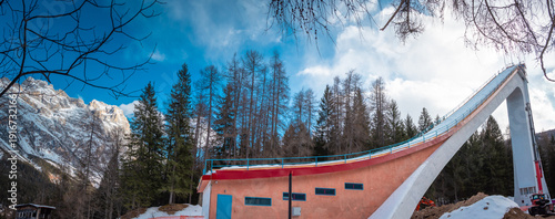 Ski jump ramp rising above alpine forest in Cortina d Ampezzo, Italy, with dramatic concrete structure, blue sky, winter light, and towering pines creating a striking mountain sports scene.