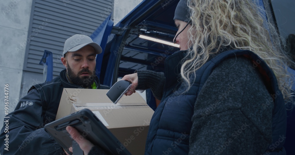 Fototapeta premium Logistics Team Unloads Parcels From Delivery Van During Snowfall. Man Moves Boxes While Woman Uses Scanner and Tablet Check Shipments. Coordinated Winter Delivery Operations in Cold Conditions.