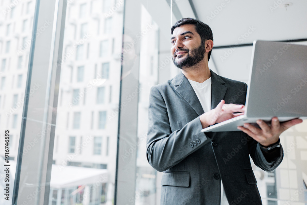 Fototapeta premium Man holds laptop while standing by a large window in a modern office space during daytime