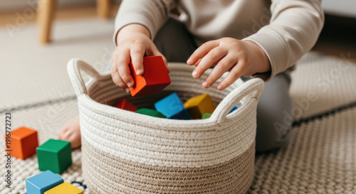 Child picking up colorful wooden block toy and putting it into a woven basket on a carpet. Concept of tidying up with easy cleanup and organization for kid.