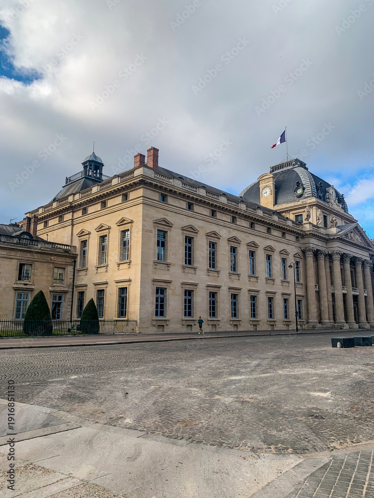 Obraz premium Majestic historic building with classical columns and a domed roof under a cloudy blue sky in Paris. Wide stone square and elegant French architecture featuring the national flag.
