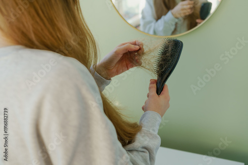 A young woman gently pulls loose hair from a hairbrush. The black brush is filled with fallen hair, emphasizing hair loss and daily hair care routine.