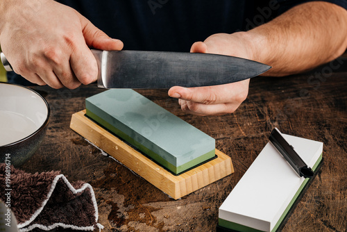 A man sharpens a knife with a grindstone on a rustic wooden table.