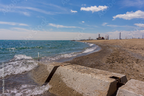 Lake Ontario, Toronto, Canada - Gentle waves roll onto a sandy shoreline beside a concrete breakwater under a bright blue sky with scattered clouds.