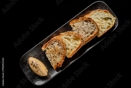 Photos Slices of crusty artisan sourdough bread and rye bread arranged on a dark plate with a quenelle of whipped butter, shot from a top-down perspective against a black background