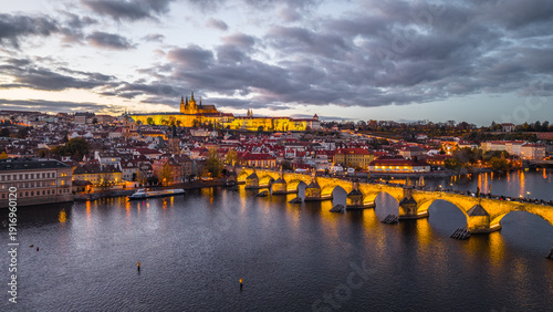 Prague Castle stands on the hill while Charles Bridge stretches over the Vltava River. The city lights glow as dusk settles, creating a scenic view of Czechia.