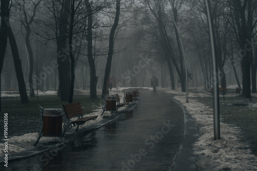 A long, wet pedestrian path curves through a foggy park during the winter thaw. Wooden benches and trash bins line the walkway, which is flanked by patches of melting snow and silhouettes of bare tree
