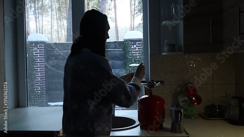 A young Ukrainian woman lights a gas stove with a match to make herself coffee during a power outage. The intimate kitchen scene highlights everyday resilience and adaptation to electricity shortages 