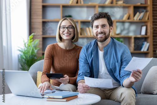 Smiling young couple sitting on a couch at home, collaborating on personal finances, calculating expenses, and filing taxes using a laptop and documents