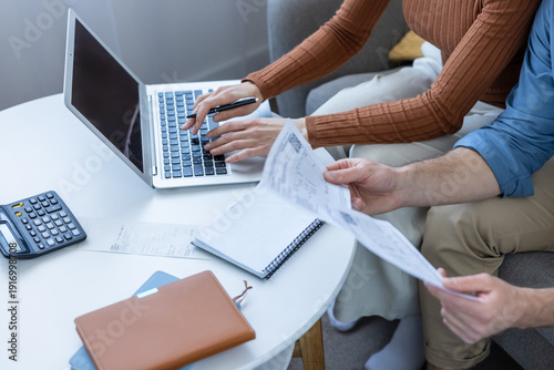 Fotografia Couple sitting on a sofa, reviewing paper documents and bills while one person t