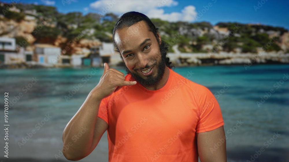 Fototapeta premium Man pointing finger at camera on sandy beach by turquoise sea wearing bright orange shirt and beard; playful confidence.