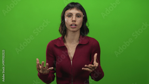 Canvas-taulu Young hispanic woman wearing round glasses and burgundy sweater gesturing hands in studio with green backdrop; communication serenity