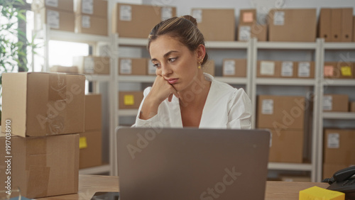 Slika na platnu Young blonde woman with hand on chin beside stacked parcel boxes and open laptop in building, checking orders and pausing at desk; fatigue