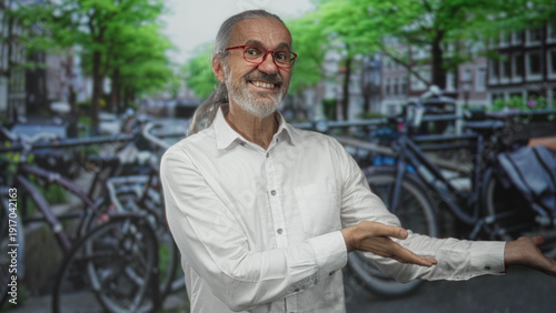 Man in white shirt with red glasses and long grey hair gesturing to parked bicycle at a street bike parking rack, smiling; community sharing welcoming.