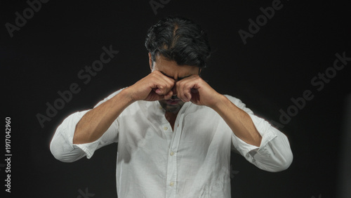 Young hispanic man rubbing his eyes in a black studio setting, wearing a white shirt and a tired expression; fatigue.