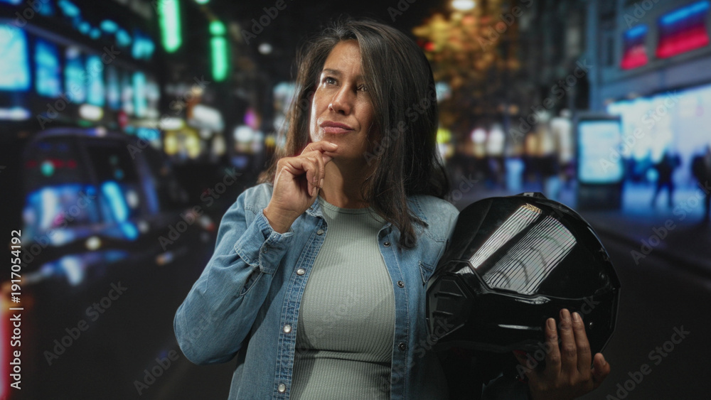 Fototapeta premium Woman middleaged brunette holding black motorcycle helmet, hand on chin in busy city street at night with neon signs and blurred pedestrians; thoughtful planning.