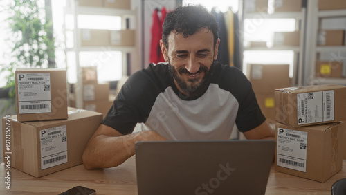 Bild auf Leinwand Man typing on laptop amid stacked packaging boxes and shipping labels in a building workspace for an ecommerce small business; entrepreneurial focused