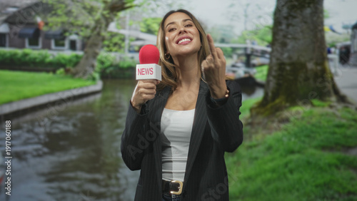 Bild auf Leinwand Woman reporter holding red microphone labeled news, wearing blazer and white top, showing middle finger on street by a canal and trees; defiant attitude