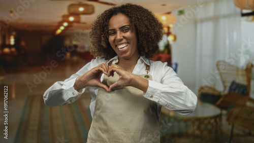 Foto Young woman baker wearing apron in hotel lobby forms heart with hands while smiling broadly and looking at camera; joy hospitality welcome