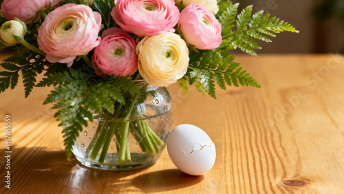 Delicate ranunculus bouquet with pink and yellow blooms and fern accents, glass vase on warm wooden surface for spring editorial