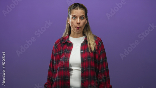 Bild auf Leinwand Young woman puckers lips showing mouth gesture wearing red plaid shirt and white tee in studio purple backdrop; playful curiosity