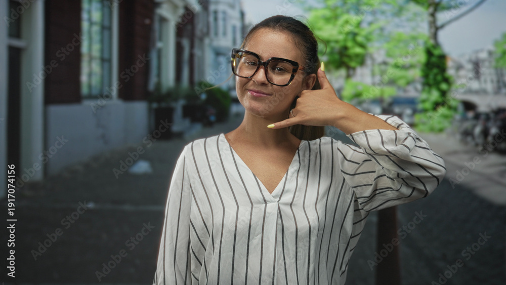 Obraz premium Woman with hand shaped like phone to ear and mouth on a city street, wearing glasses and striped blouse, smiling with closed eyes; playful moment.