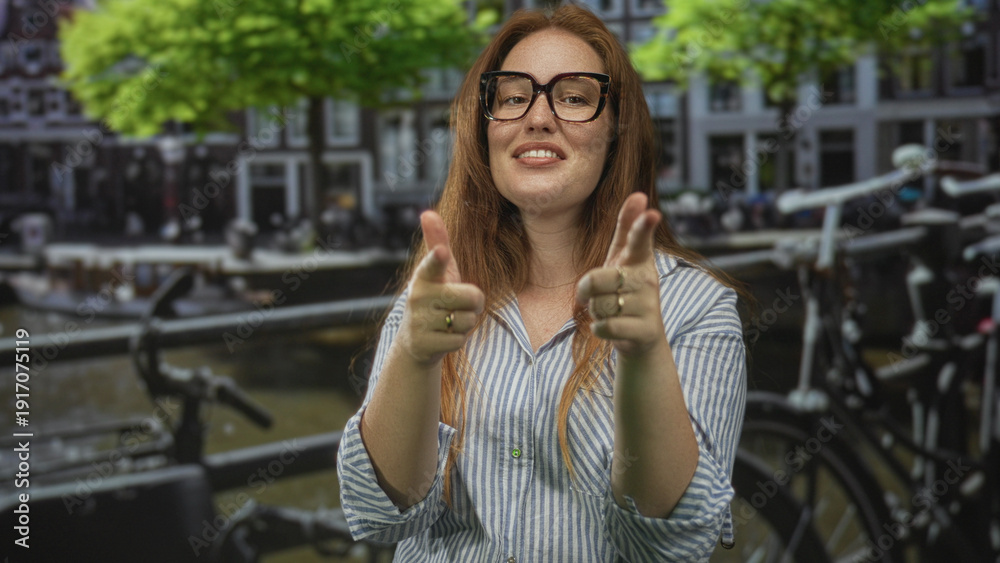 Fototapeta premium Woman wearing glasses and striped shirt points fingers to camera on street beside canal boats; playfulness.