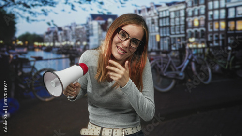 Canvastavla Woman redhead holding megaphone while covering mouth with hand on canal street lined with bicycles and buildings; playful protest