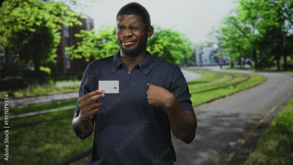 Fototapeta premium Man holding a credit card in left hand and points finger to card near his chest on a tree lined urban street with tram tracks; doubt identity security.