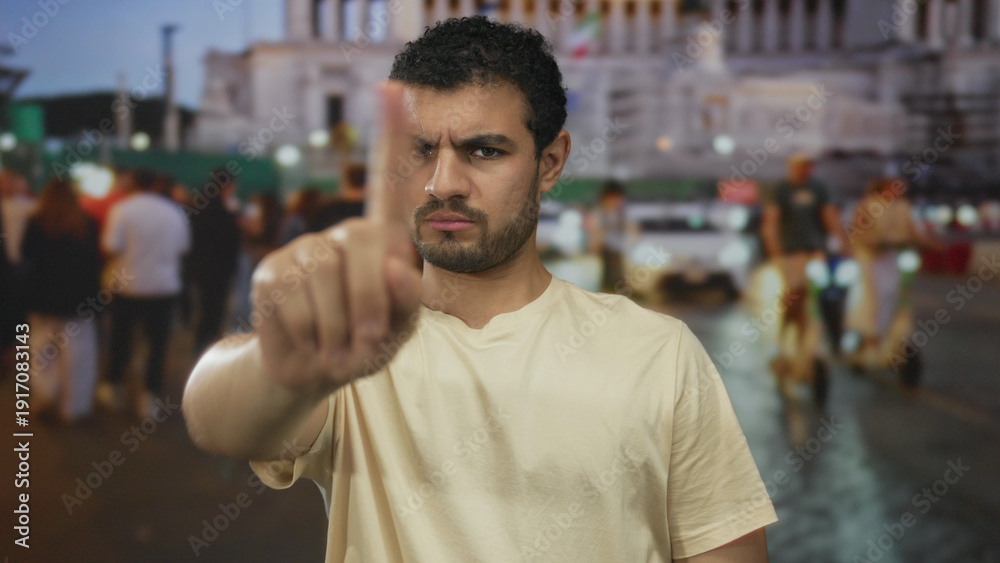 Fototapeta premium Young hispanic man pointing index finger directly at camera in a crowded urban street at dusk; serious.