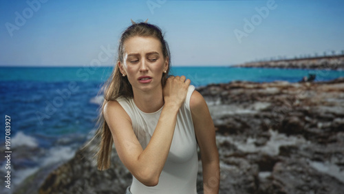 фотографія Woman holding her shoulder and grimacing, showing shoulder pain on rocky seaside near a building pier under blue sky; pain