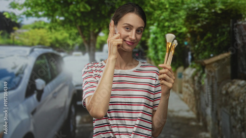 Fotografi Woman holding makeup brushes points finger to cheek in street near parked car and stone wall; confidence self care
