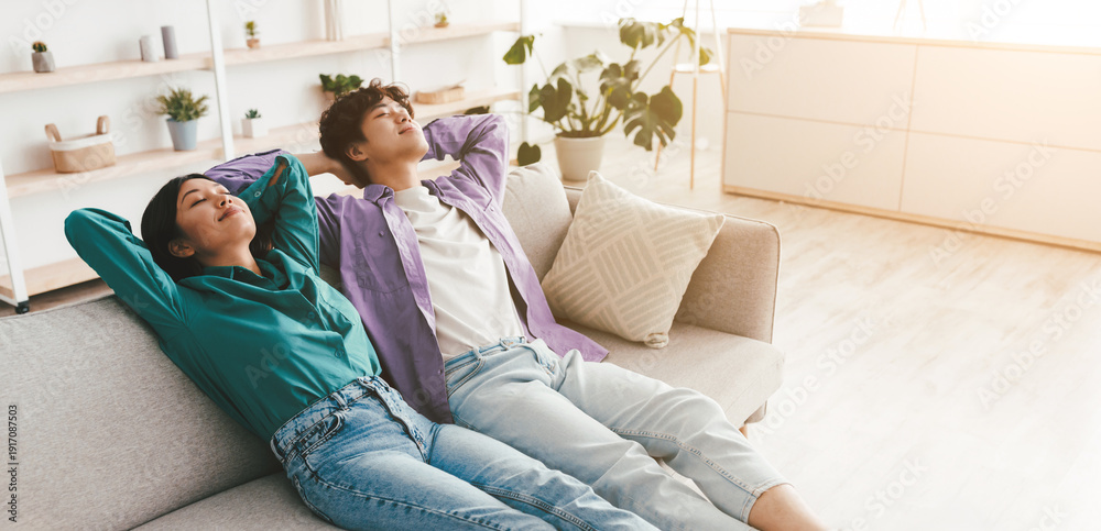 Obraz premium Calm young Asian couple relaxing on couch with their hands behind heads at home, blank space. Carefree girlfriend and boyfriend chilling on sofa, enjoying peaceful weekend morning