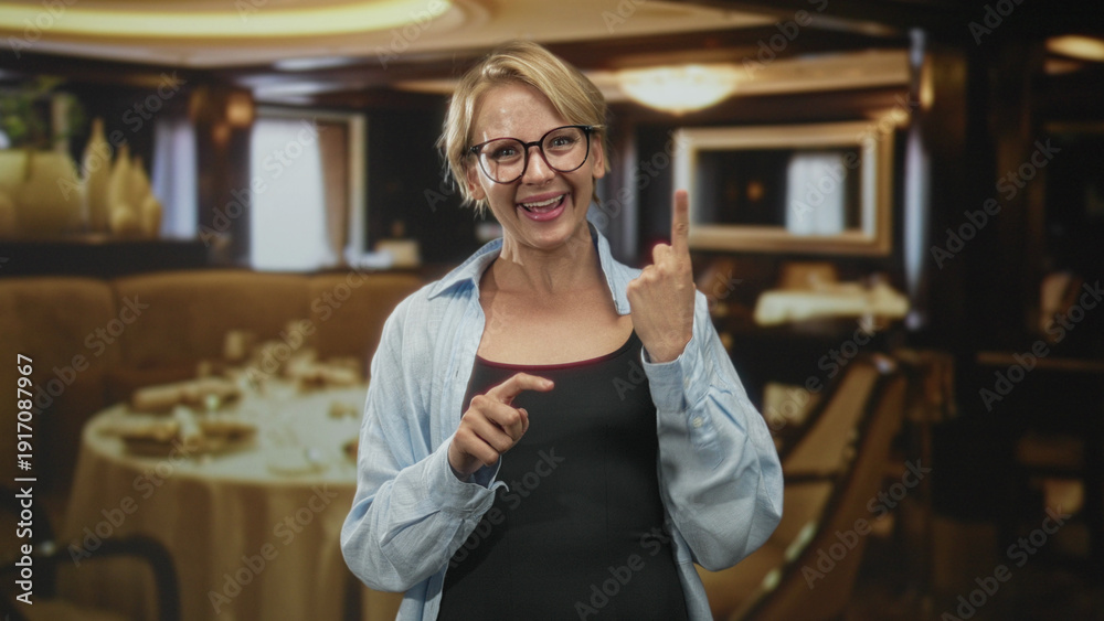 Fototapeta premium Blond woman smiling with glasses, eyes closed, crossing fingers and pointing with hands while wearing casual shirt in restaurant; hopeful.